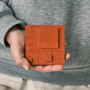 Könnte beinhalten: Ein quadratisches Objekt in Terrakotta-Farbe mit einem Rastermuster und erhabenen quadratischen Elementen. Das Objekt wird in der Hand einer Person gehalten, mit einem grauen Sweatshirt im Hintergrund. Es sieht aus wie ein Puzzle oder eine dekorative Fliese.