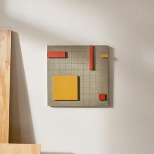 May include: A geometric wall art piece featuring a gray grid background with various colored shapes. The shapes include a large yellow square, red rectangles, and a small orange square. The artwork is mounted on a white wall.