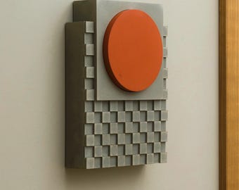 May include: A geometric wall sculpture featuring a gray rectangular base with a checkerboard pattern. A gray square supports an orange circle. The sculpture is mounted on a light gray wall.