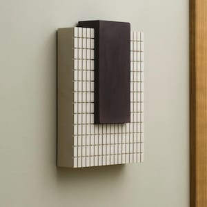 Puede incluir: Una obra de arte rectangular montada en la pared con un patrón de cuadrícula y un bloque marrón oscuro en la parte superior. La cuadrícula está compuesta por pequeños rectángulos verticales de color crema. El bloque marrón oscuro es sólido y rectangular.