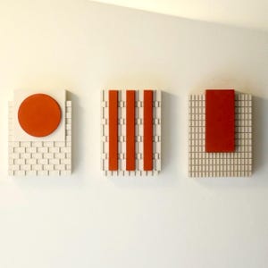 Könnte beinhalten: Drei abstrakte Wandkunstwerke. Jedes hat ein geometrisches Design mit roten und cremefarbenen Elementen. Eines hat einen roten Kreis, ein anderes drei vertikale rote Balken und das dritte ein rotes Rechteck.