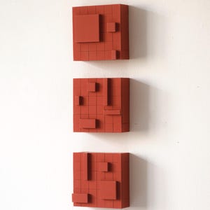 Puede incluir: Tres obras de arte murales geométricas rojas. Cada pieza es un cuadrado con un patrón de cuadrícula y varias formas rectangulares y cuadradas adjuntas. El arte está montado en una pared blanca, creando una estética moderna y minimalista.