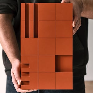 May include: A terracotta-coloured rectangular block with a grid pattern and cut-out sections. The block is held by a person and features a series of vertical and horizontal rectangular shapes, creating a geometric design. The overall aesthetic is modern and minimalist.