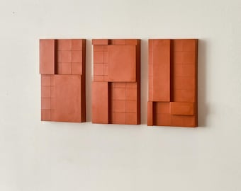 May include: Three terracotta-colored wall art panels with geometric designs. Each panel has a different arrangement of rectangular and square shapes, some with grid patterns. The panels are mounted on a white wall, with a wooden table in the foreground.