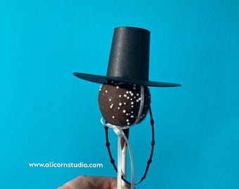 May include: A chocolate-covered cake pop decorated with white sprinkles, a black top hat, and a white ribbon. The cake pop is on a stick and has a black string detail. The website address www.alicornstudio.com is visible.