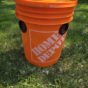 May include: An orange Home Depot bucket with a matching lid. The bucket has black vents and the Home Depot logo in white. The bucket is sitting on green grass.