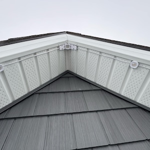 May include: Exterior view of a roof's corner, featuring gray shingles and white siding with a perforated pattern. White lights are mounted on the siding. The sky is overcast.