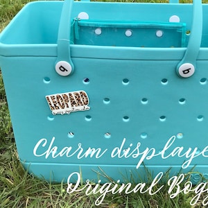 May include: A turquoise blue tote bag with a white handle and white polka dot zippered pouch inside. The bag has a white letter "B" on the side and a leopard print charm with the text "Leopard" and "My signature color" on the front. The bag is sitting on a bed of green grass. Charm displayed on Original  Bogg.