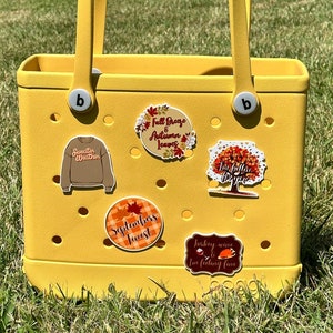 May include: A yellow tote bag with two handles and several decorative charms. The charms include a brown sweater, fall leaves, a tree, and the text "September's Finest" and "Turkey wine & fall feeling fine".