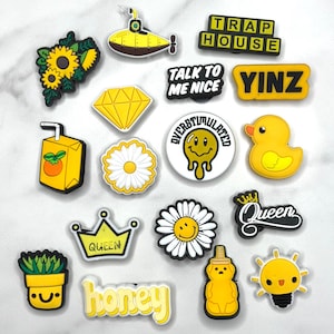 May include: A collection of yellow and black shoe charms. Designs include a sunflower, a submarine, a diamond, a daisy, a rubber duck, a crown with the word "Queen", a honey jar, and the words "Trap House", "Talk to me nice", "Yinz", "Overstimulated", and "honey".