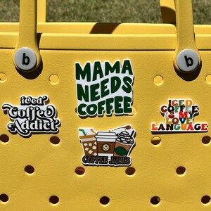 K&ouml;nnte beinhalten: Gelbe Tragetasche mit verschiedenen Kaffee-Aufklebern. Die Aufkleber zeigen "Mama Needs Coffee", "Iced Coffee is My Love Language", "Coffee Addict" und "Coffee Vibes".