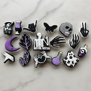 May include: A set of 15 black and white charms with a spooky Halloween theme. The charms include a skull, a ghost, a skeleton, a potion bottle, a crescent moon, a leaf, a crystal, a hand, and a skull with a peace sign. The charms are made of silicone and are perfect for adding a touch of Halloween to your shoes, bags, or other accessories.