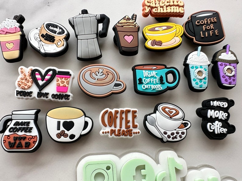 Op de afbeelding: Een verzameling kleurrijke koffie-thema charms met verschillende ontwerpen en spreuken, waaronder "Peace Love Coffee", "Make Coffee Not War", "Coffee For Life", "Drink Coffee Outdoors" en "I Need More Coffee".