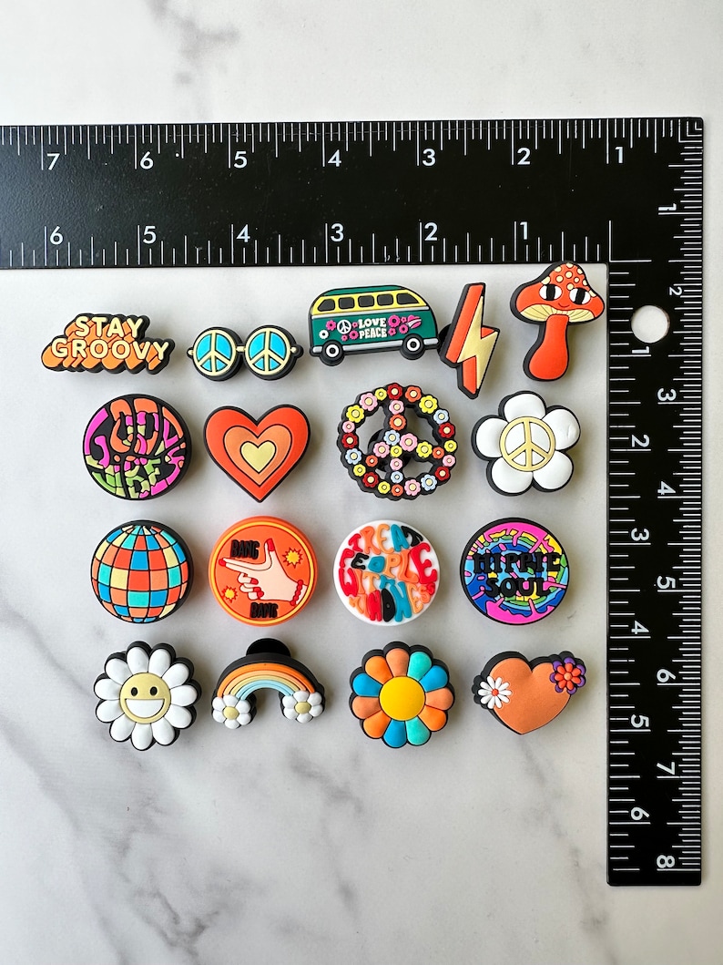 May include: A set of 16 colourful, groovy, and peace-themed shoe charms. The charms feature various designs, including peace signs, flowers, hearts, rainbows, and a smiley face. The charms are made of rubber and have a glossy finish.