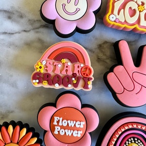 May include: A set of nine colorful, retro-style flower and rainbow-themed pins. The pins feature peace signs, smiley faces, and the text "Stay Groovy" and "Flower Power".