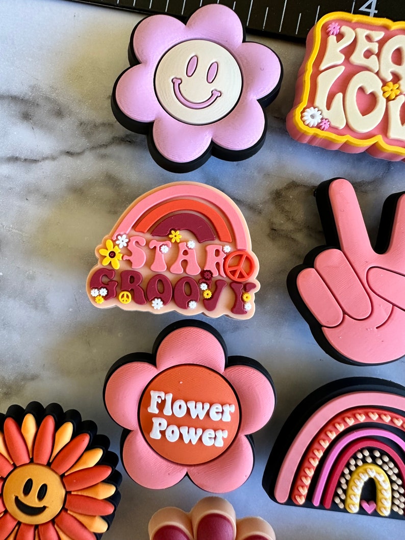 May include: A set of nine colorful, retro-style flower and rainbow-themed pins. The pins feature peace signs, smiley faces, and the text "Stay Groovy" and "Flower Power".