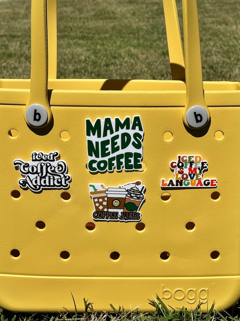 K&ouml;nnte beinhalten: Gelbe Tragetasche mit verschiedenen Kaffee-Aufklebern. Die Aufkleber zeigen "Mama Needs Coffee", "Iced Coffee is My Love Language", "Coffee Addict" und "Coffee Vibes".