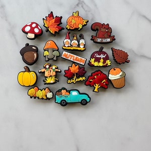 Op de afbeelding: Een verzameling van 16 herfst-thema schoencharms met pompoenen, bladeren, kabouters, een eekhoorn, een vrachtwagen en een vogelverschrikker. De charms zijn gemaakt van kleurrijk rubber en hebben verschillende herfstontwerpen.