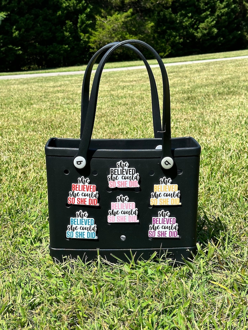 May include: A black tote bag with a white handle and multiple colorful stickers that say "She Believed She Could So She Did."