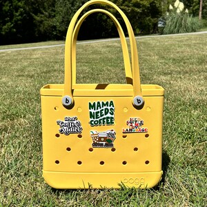 K&ouml;nnte beinhalten: Gelbe Tragetasche mit verschiedenen bunten Aufklebern, darunter "Mama Needs Coffee", "Iced Coffee Is My Love Language" und "Coffee Junkie". Die Tasche ist von der Marke "BOGG"