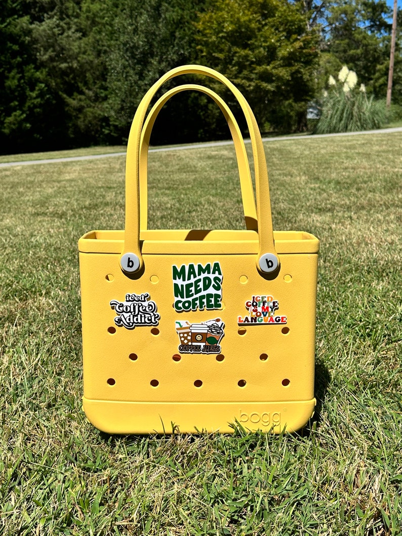 K&ouml;nnte beinhalten: Gelbe Tragetasche mit verschiedenen bunten Aufklebern, darunter "Mama Needs Coffee", "Iced Coffee Is My Love Language" und "Coffee Junkie". Die Tasche ist von der Marke "BOGG"
