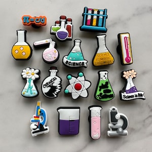 May include: A set of 17 colorful science-themed shoe charms. The charms feature beakers, test tubes, microscopes, and other science-related imagery. Some charms have text, such as "Science is magic that works" and "Science is lit".