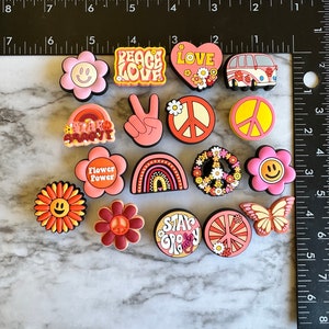 May include: A collection of 14 colorful, peace-themed shoe charms. The charms feature peace signs, flowers, smiley faces, rainbows, and the words "Peace Love", "Star Groovy", "Flower Power", and "Stay Groovy".