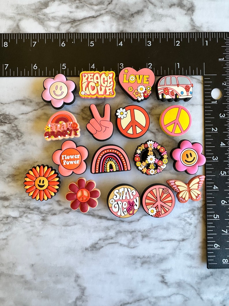 May include: A collection of 14 colorful, peace-themed shoe charms. The charms feature peace signs, flowers, smiley faces, rainbows, and the words "Peace Love", "Star Groovy", "Flower Power", and "Stay Groovy".