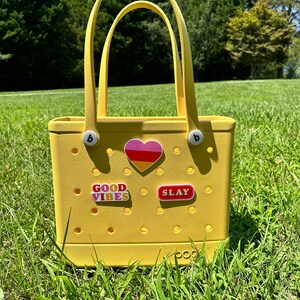 Op de afbeelding: Een gele tote bag met een hart, "GOOD VIBES" en "SLAY" stickers. De tas heeft een wit logo met de tekst "boo" aan de onderkant.
