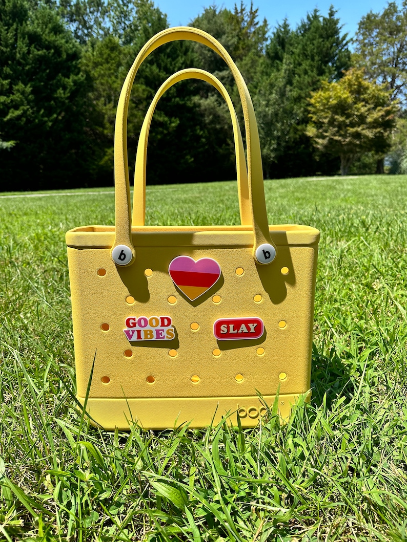 Op de afbeelding: Een gele tote bag met een hart, "GOOD VIBES" en "SLAY" stickers. De tas heeft een wit logo met de tekst "boo" aan de onderkant.