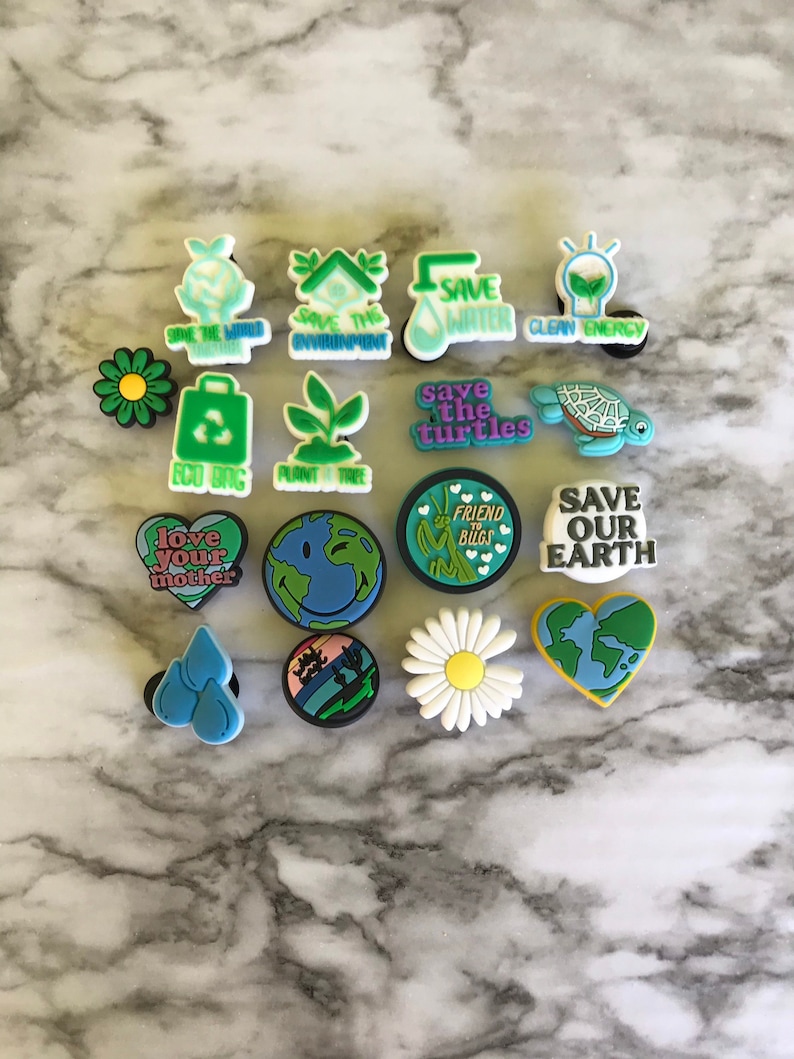 May include: A set of 15 colorful shoe charms with environmental themes. The charms feature images of a lightbulb, a plant, a turtle, a heart, a daisy, a water droplet, a globe, and a recycling bin. The charms also include text such as "Save the World", "Save the Environment", "Save Water", "Clean Energy", "Save the Turtles", "Eco Bag", "Plant a Tree", "Love Your Mother", "Friend Bugs", and "Save Our Earth".