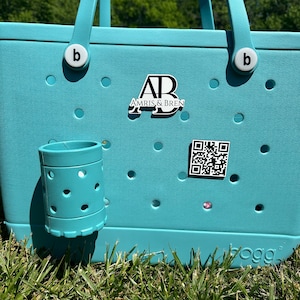 May include: A light blue tote bag with a white "b" on each handle. The bag has a white and black logo that says "AB Amris & Bren" and a black and white QR code. The bag is sitting on green grass.