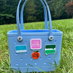 May include: A light blue tote bag with a variety of colorful stickers. The stickers include phrases like "Your words matter", "Just Speechy", and "Hello I'm the Speech Pathologist".