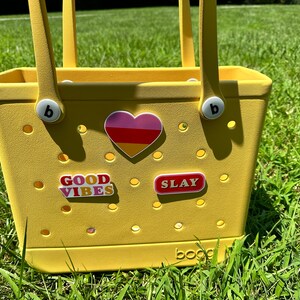 Op de afbeelding: Gele tote bag met een hart, "GOOD VIBES" en "SLAY" stickers. De tas heeft een witte "b" op elke handgreep.