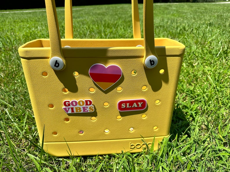 Op de afbeelding: Gele tote bag met een hart, "GOOD VIBES" en "SLAY" stickers. De tas heeft een witte "b" op elke handgreep.