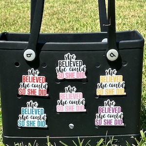 May include: A black tote bag with six colorful stickers that say "She Believed She Could So She Did." The stickers are in different colors, including pink, red, blue, yellow, and purple.