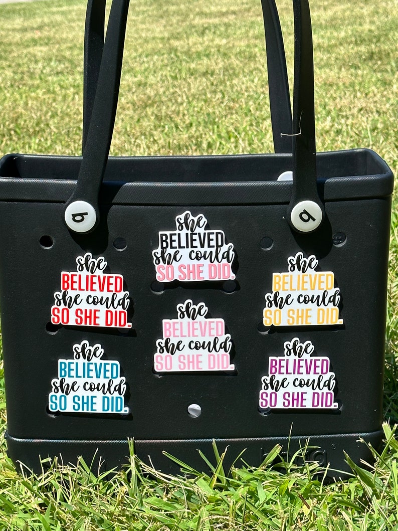 May include: A black tote bag with six colorful stickers that say "She Believed She Could So She Did." The stickers are in different colors, including pink, red, blue, yellow, and purple.