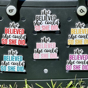 May include: Seven colorful stickers with the text "She believed she could so she did." The stickers are on a black surface.