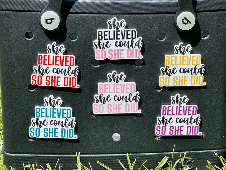 May include: Seven colorful stickers with the text "She believed she could so she did." The stickers are on a black surface.