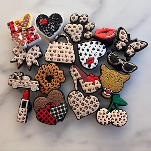 May include: A collection of 16 colorful shoe charms featuring various designs, including a leopard print purse, a red heart, a red lip, a leopard print shoe, a leopard print bow, a leopard print cherry, a leopard print truck, a leopard print paw print, a leopard print butterfly, a leopard print cheetah, a leopard print flower, a leopard print heart, a leopard print lipstick, a leopard print sunglasses, a leopard print love sign, and a black and red plaid heart.