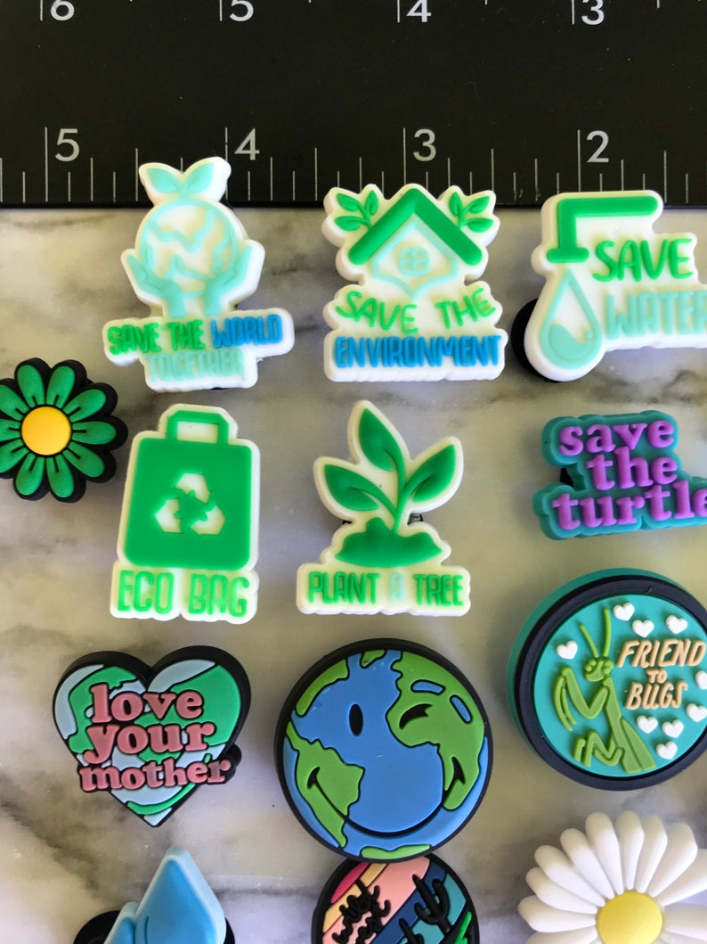 May include: A collection of 12 colorful, eco-friendly charm buttons featuring various designs, including a green eco bag, a plant, a daisy, a heart with the text "love your mother", a smiling earth, a praying mantis with the text "friend to bugs", a house with the text "save the environment", a water drop with the text "save water", a turtle with the text "save the turtle", and a hand holding a globe with the text "save the world together".
