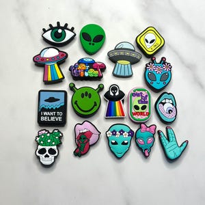 May include: A collection of 18 colorful alien-themed shoe charms. The charms feature various designs, including aliens, UFOs, eyes, smiley faces, and the text "I want to believe".