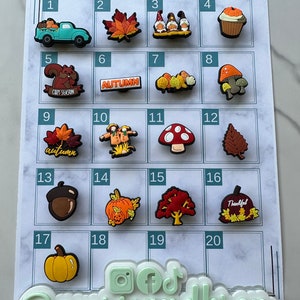 Op de afbeelding: Een kaart met 20 genummerde vakjes, elk met een andere herfst-thema charme. De charmes omvatten een vrachtwagen, bladeren, kabouters, een cupcake, een eekhoorn, paddenstoelen, een eikel, een pompoen, een boom en een dankbare pompoen. De kaart is getiteld "Amris & Bren" en heeft social media-iconen onderaan.