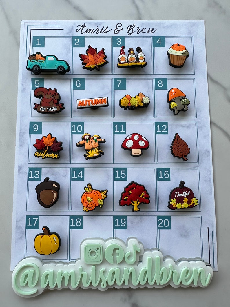 Op de afbeelding: Een kaart met 20 genummerde vakjes, elk met een andere herfst-thema charme. De charmes omvatten een vrachtwagen, bladeren, kabouters, een cupcake, een eekhoorn, paddenstoelen, een eikel, een pompoen, een boom en een dankbare pompoen. De kaart is getiteld "Amris & Bren" en heeft social media-iconen onderaan.