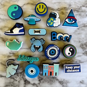 May include: A collection of 15 colorful shoe charms featuring various designs, including a smiley face, a UFO, an astronaut, an evil eye, a yin yang symbol, a band-aid, a teapot, a crab, a building, a cat, and a phrase that says "adore you".