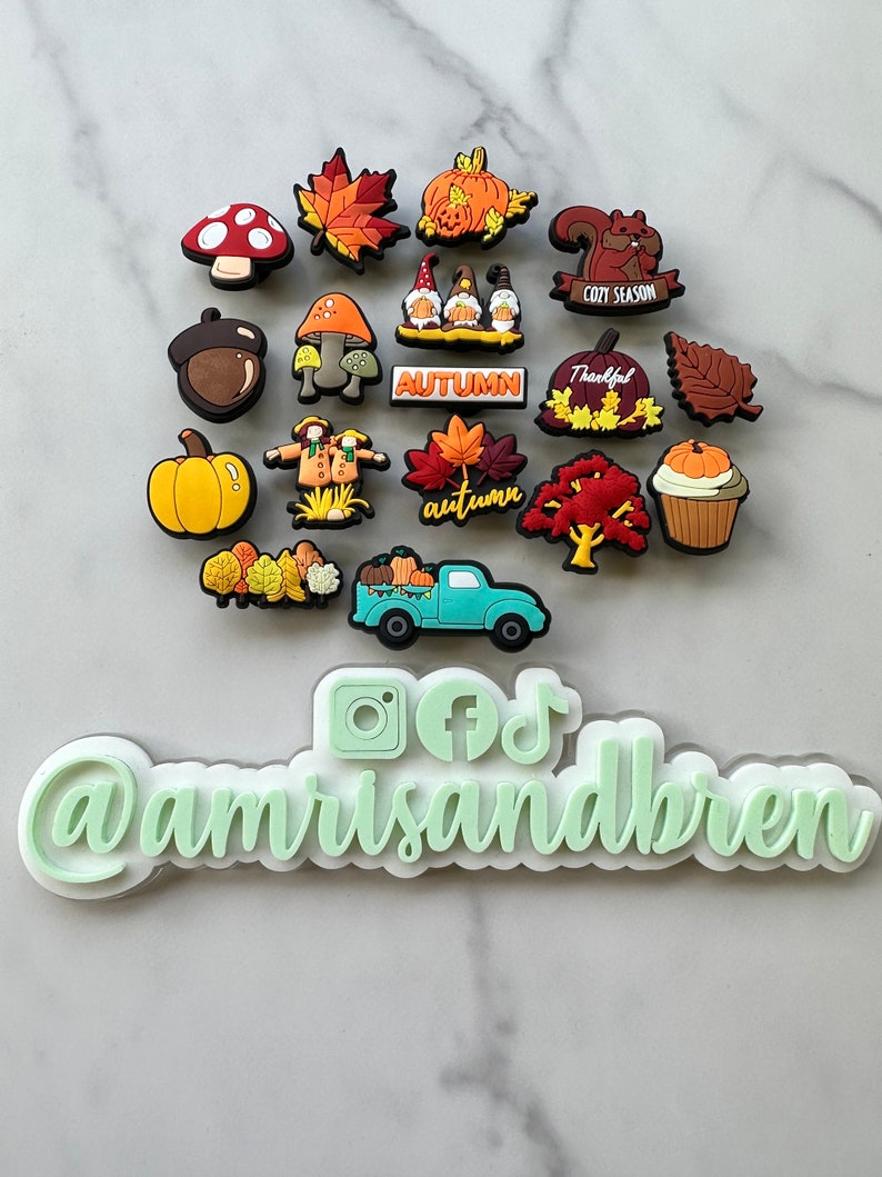 Op de afbeelding: Een verzameling van 18 kleurrijke herfst-schoenbedels, waaronder pompoenen, bladeren, paddenstoelen, kabouters, een vrachtwagen en een eekhoorn. De bedels zijn in een cirkel op een wit marmeren oppervlak gerangschikt. De tekst "@amrisandbren" is zichtbaar onder de bedels.