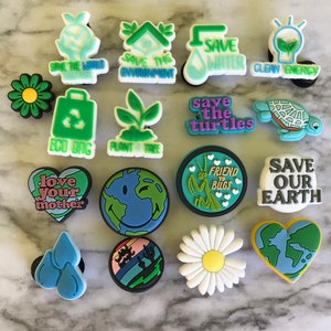 May include: A collection of 15 colorful shoe charms with various environmental themes. The charms feature images of a house, a lightbulb, a turtle, a daisy, a heart, a bug, a plant, a world, a bag, and water drops. The charms also include text such as "Save the World", "Save the Environment", "Save Water", "Clean Energy", "Save the Turtles", "Friend to Bugs", "Save Our Earth", "Love Your Mother", "Eco Bag", and "Plant a Tree".