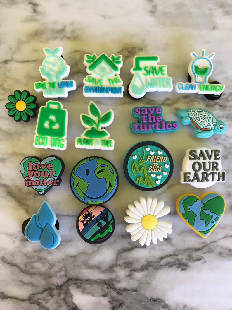 May include: A collection of 15 colorful shoe charms with various environmental themes. The charms feature images of a house, a lightbulb, a turtle, a daisy, a heart, a bug, a plant, a world, a bag, and water drops. The charms also include text such as "Save the World", "Save the Environment", "Save Water", "Clean Energy", "Save the Turtles", "Friend to Bugs", "Save Our Earth", "Love Your Mother", "Eco Bag", and "Plant a Tree".