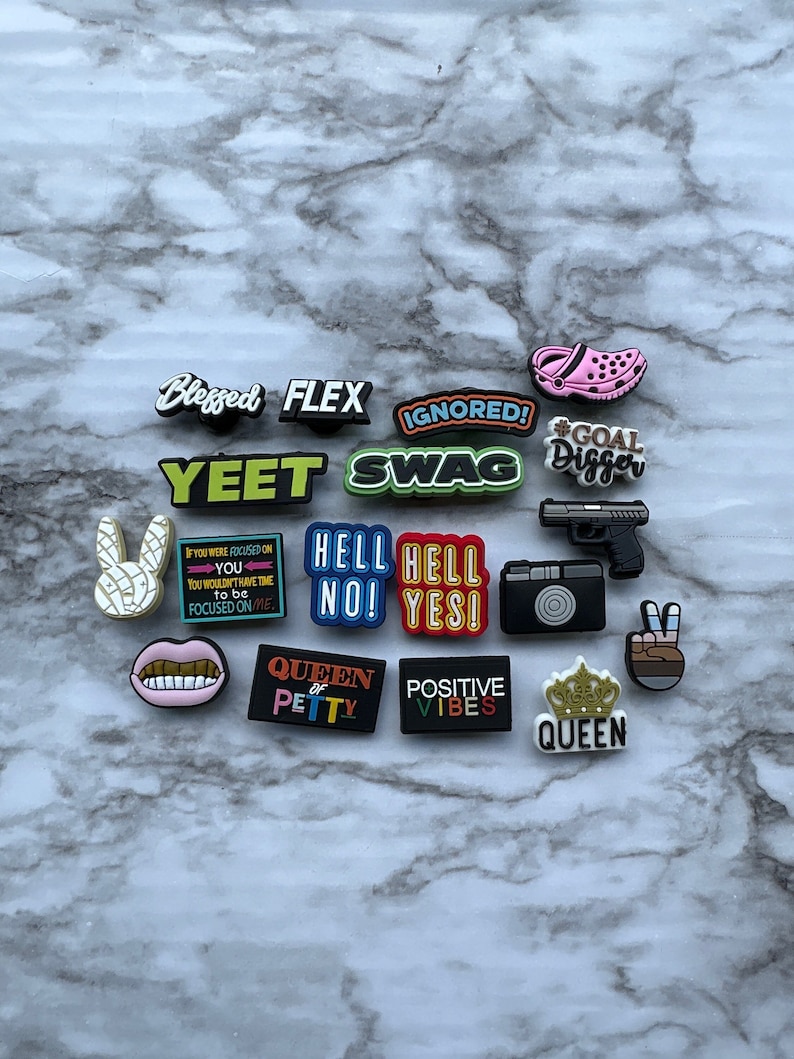 May include: A collection of colorful shoe charms with various phrases and designs, including "Blessed", "Flex", "Yeet", "Swag", "Ignored", "Goal Digger", "Hell No", "Hell Yes", "Queen of Petty", "Positive Vibes", and "Queen".