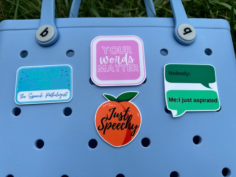 K&ouml;nnte beinhalten: Eine blaue Tragetasche mit f&uuml;nf bunten Aufklebern. Die Aufkleber sagen "Hello I'm The Speech Pathologist", "Your Words Matter", "Just Speechy", "Nobody:" und "Me: I just aspirated".